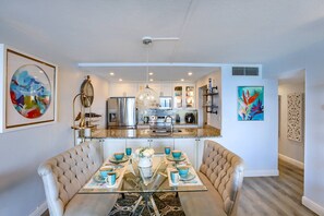 A modern kitchen with stainless steel appliances, a dining area with a glass table and cushioned chairs, and artwork on the walls.