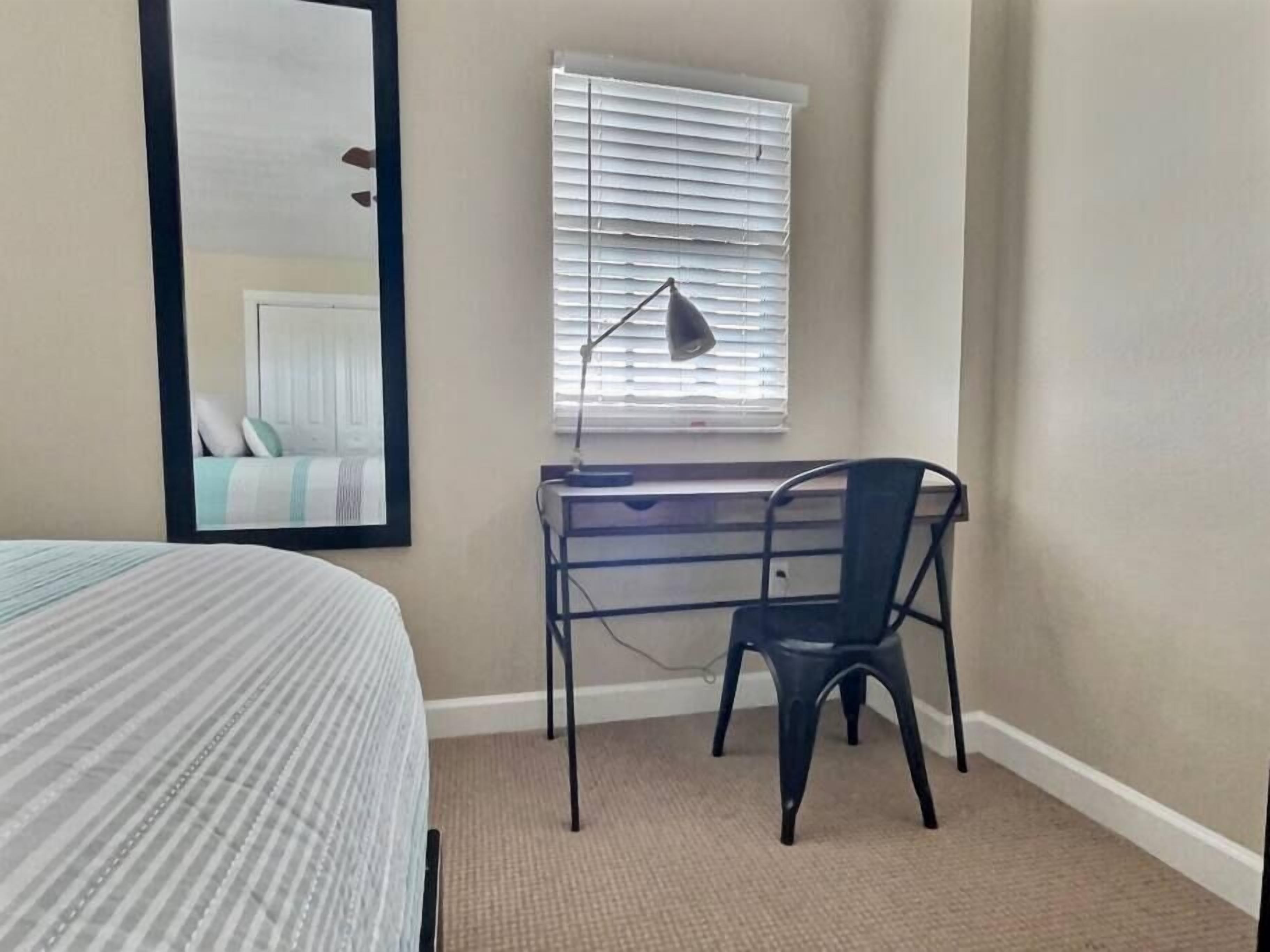Een slaapkamer met een bed, een bureau met stoel, een raam met jaloezieën en een spiegel.
