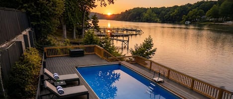 A swimming pool with lounge chairs, a dock, and a sunset view over a lake.