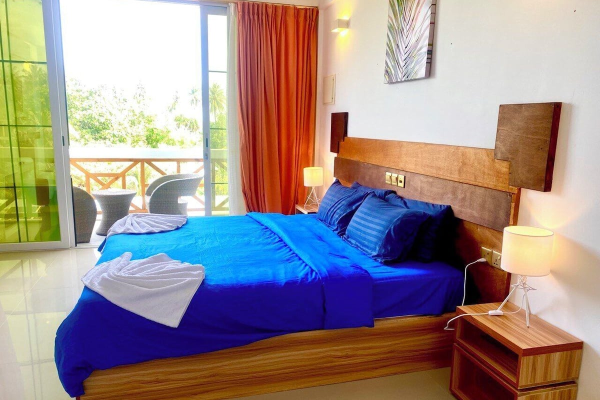 A bedroom with a large bed, a wooden headboard, a nightstand with a lamp, and a view of the outdoors through a sliding glass door.
