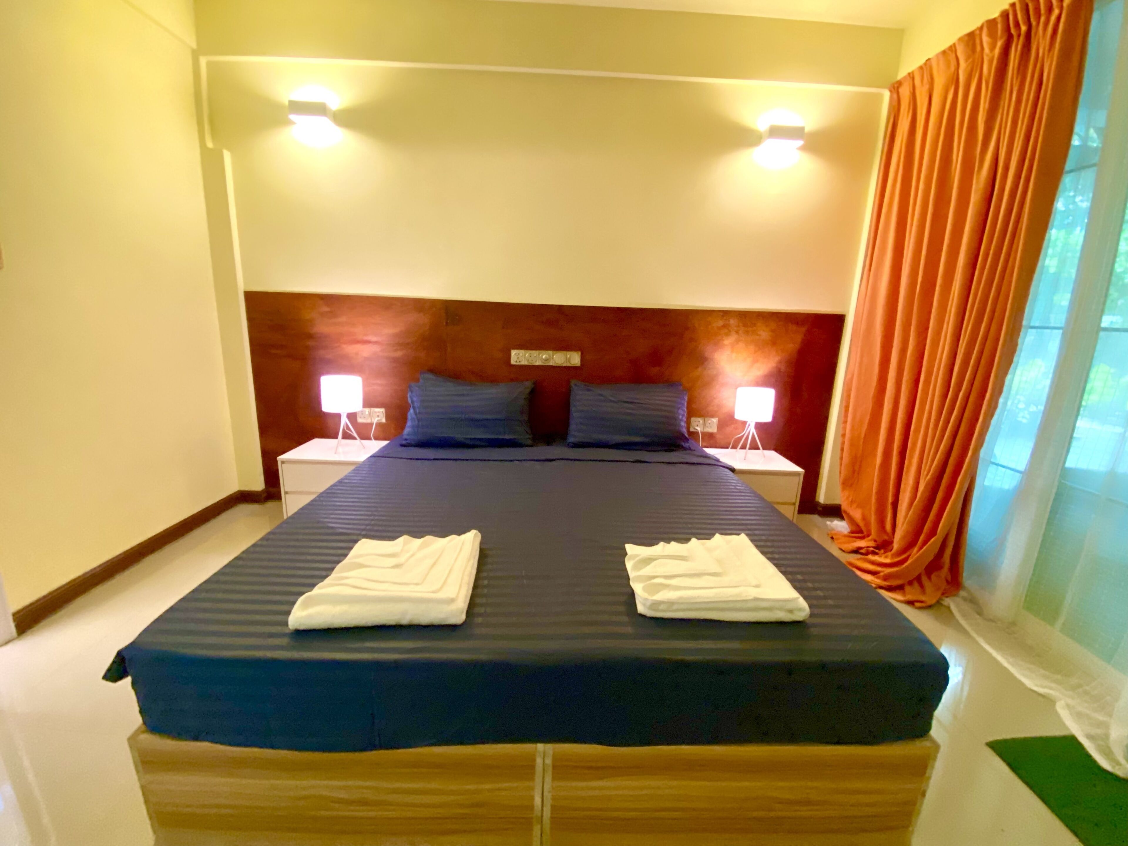 Een hotelkamer met een groot bed, twee nachtkastjes met lampen, een houten hoofdeinde en een raam met gordijnen.