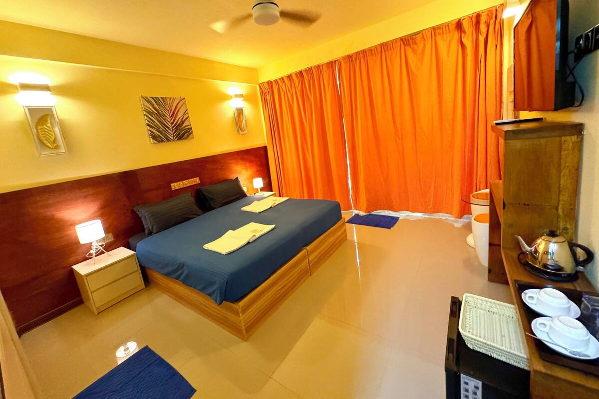 A hotel room with a bed, bedside table, lamp, curtains, a television, and a desk with a telephone and teapot.