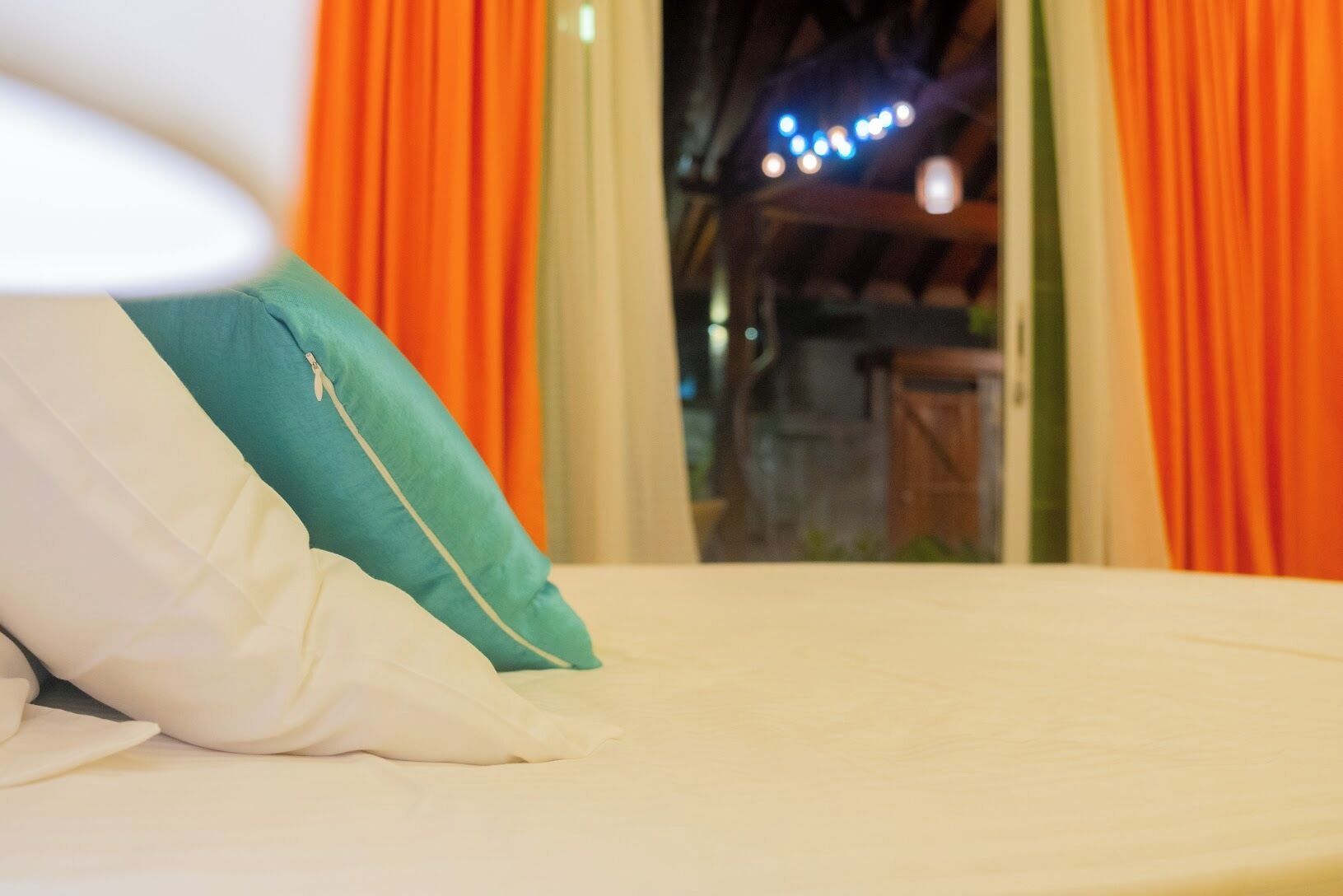 Una cama con almohadas blancas y turquesa, una ventana con cortinas naranjas, y vistas a un edificio iluminado.