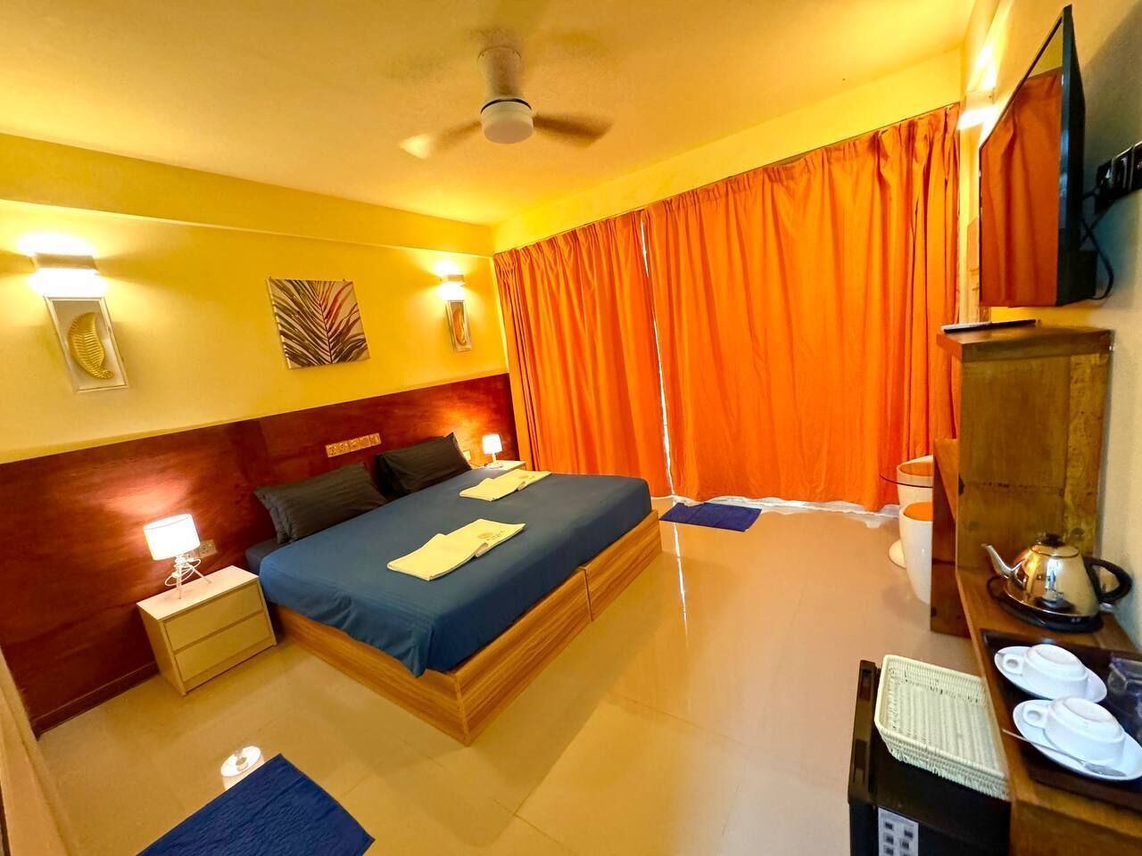 Quarto de hotel com cama, mesa de cabeceira, luminária, cortinas, televisão e uma pequena mesa com bule e xícaras.
