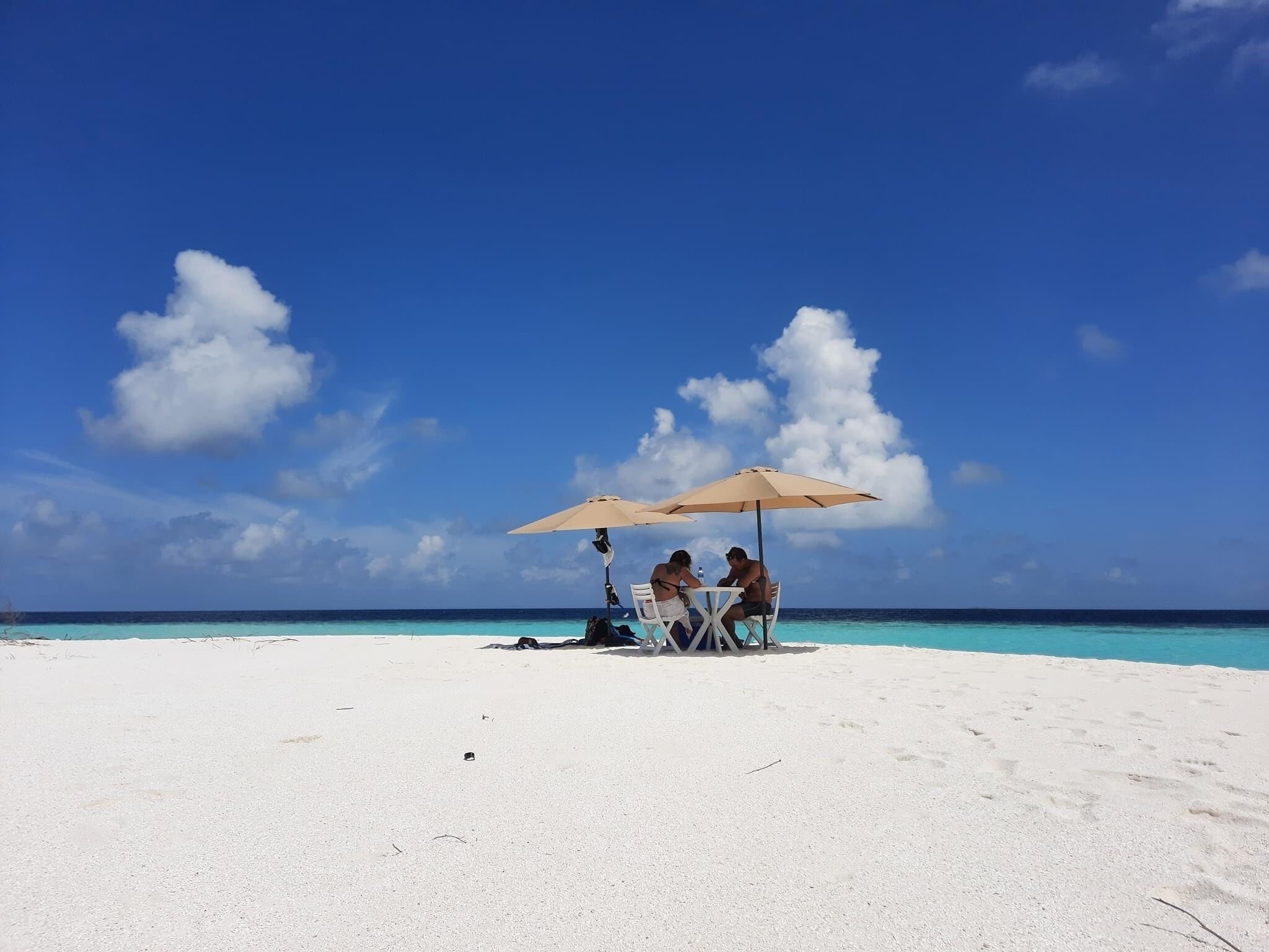 A beach scene with a clear sky, three people sitting under umbrellas, and a sandy beach.