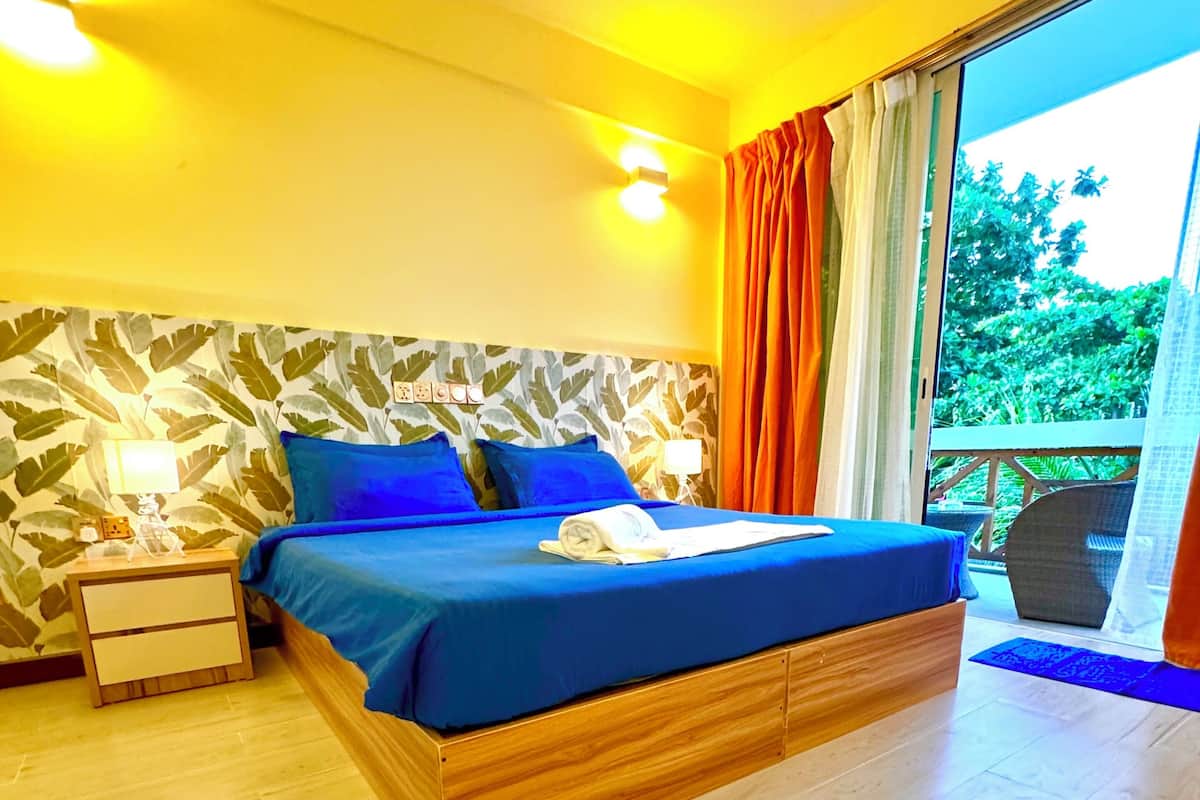 A bedroom with a wooden bed, a nightstand, a wall with leaf-pattern wallpaper, and a view of greenery through a window.
