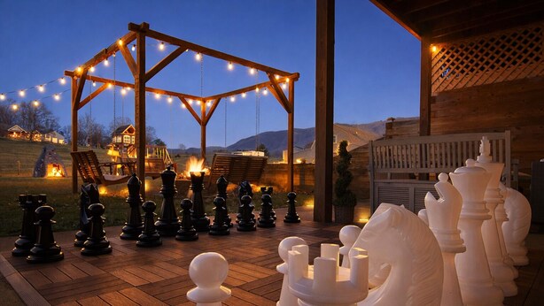 Un jeu d’échecs installé sur une terrasse en bois, surmontée d’une pergola en bois, le tout entouré d’un paysage rural au crépuscule.