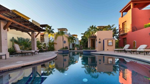 A swimming pool with lounge chairs and palm trees, surrounded by buildings with Mediterranean architecture. - Casa Patricia by Casago (Loreto)