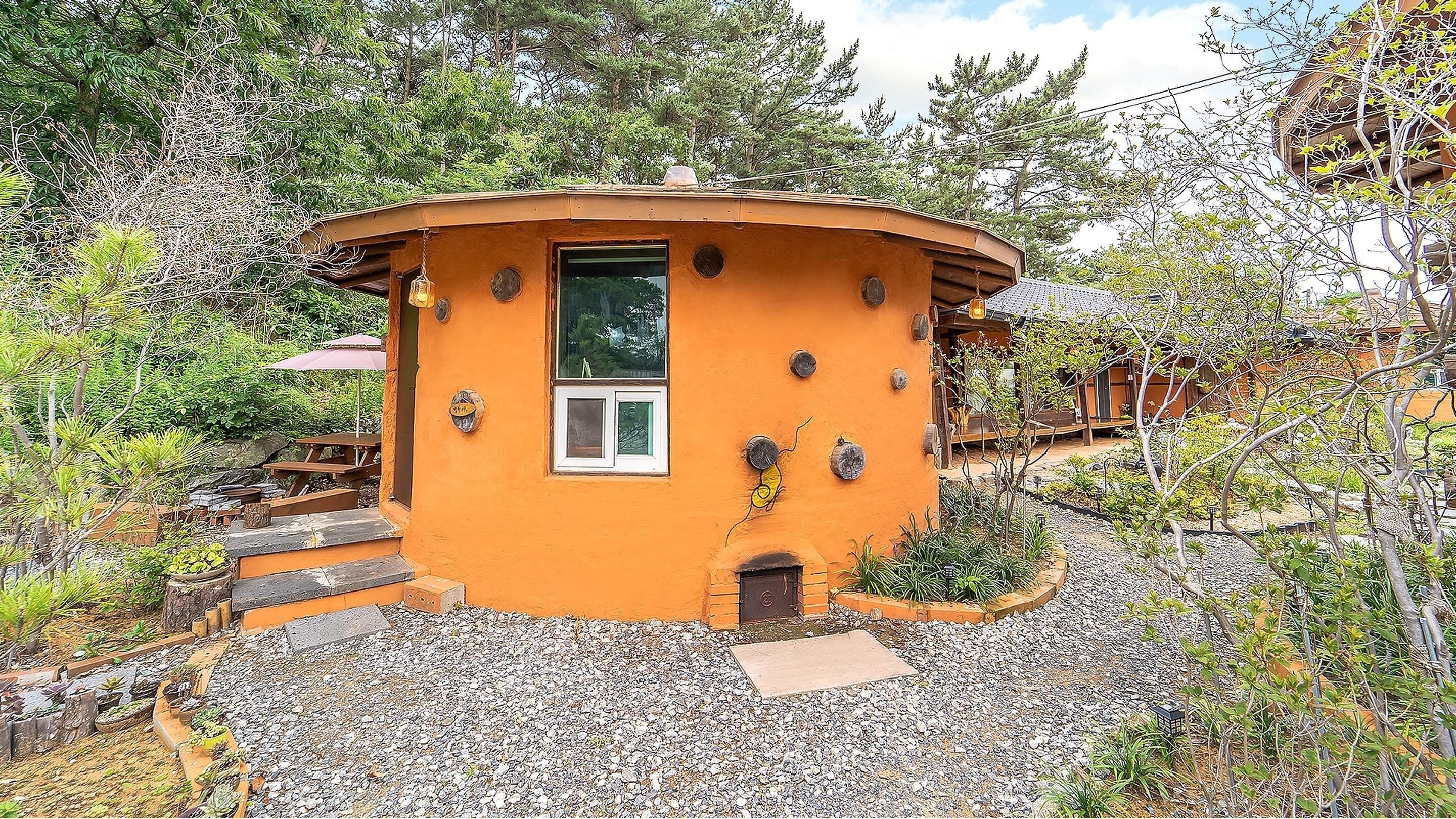 A small, orange, octagonal building with a window and a door, surrounded by gravel and plants.