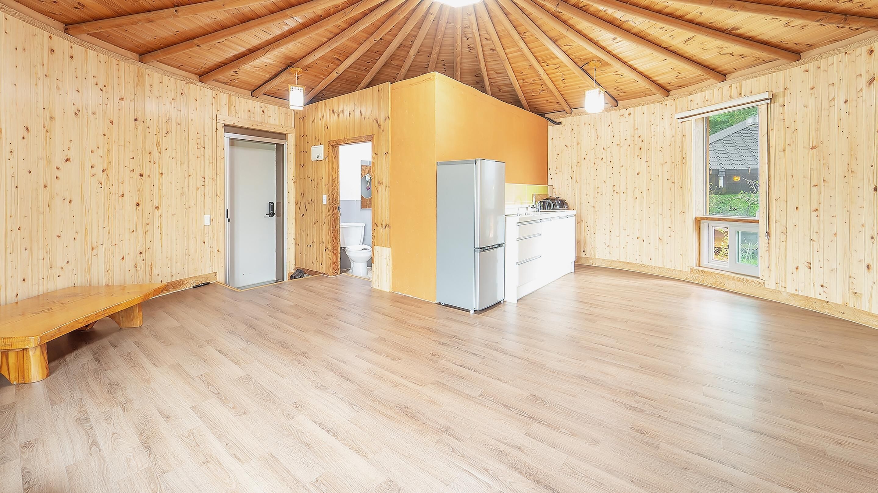 Una habitación amplia con paredes de madera y techo inclinado, un banco, una zona de cocina con refrigerador y una ventana con vistas.