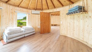 Interior de una cabaña de madera con una cama, un banco y vistas al verdor exterior.