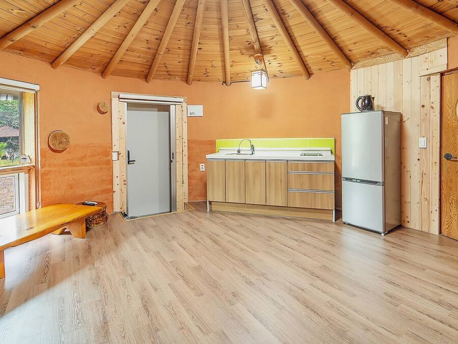 A compact kitchen with wooden cabinets, a sink, and a refrigerator. There is a bench and a door leading to another room.