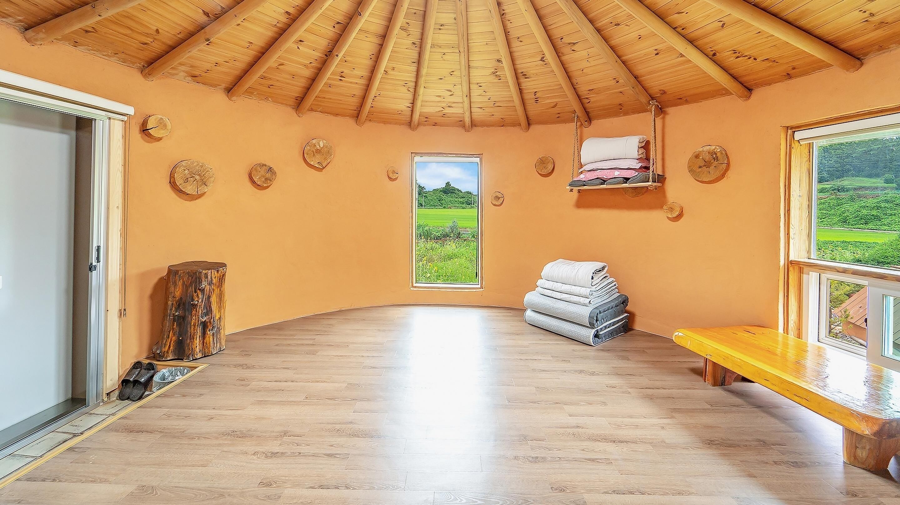 A room with a wooden ceiling, a large window with a view of a green landscape, a wooden bench, and a small shelf with folded towels.