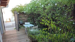 Une terrasse en bois mène à un espace patio aménagé avec une table et des chaises, le tout entouré d’une végétation luxuriante et d’une clôture en bois.