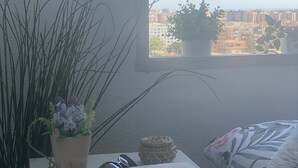 A bedroom with a window view of a cityscape, a bedside table with a book, a remote control, and a potted plant. - Casa Angel Malaga (Málaga)