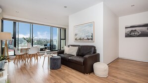 A modern living room with a leather sofa, a round coffee table, and a dining area with a white table and chairs. Large windows offer a view of the city. - Aqua Views of the bay in Mornington (Mornington)
