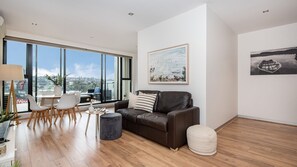 A modern living room with a leather sofa, a round coffee table, and a dining area with a white table and chairs. Large windows offer a view of the city. - Aqua Views of the bay in Mornington (Mornington)