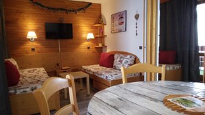 Una habitación acogedora con paredes de madera, una mesa de madera, dos sofás con cojines estampados, un televisor montado en la pared y una ventana con cortinas.