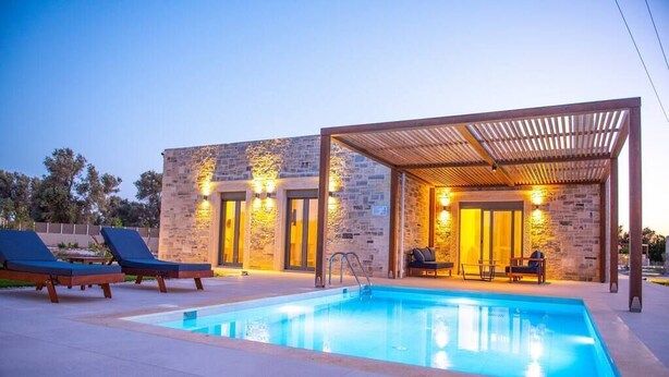 Un área moderna junto a la piscina con una piscina rectangular, sillas de descanso y una zona de estar cubierta con paredes de piedra e iluminación cálida.