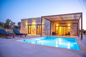 Ein moderner Poolbereich mit rechteckigem Pool, Liegestühlen und einer überdachten Sitzgruppe mit Steinmauern und warmer Beleuchtung.
