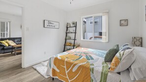 Une chambre à coucher comprenant un lit, une table de chevet, une fenêtre, une étagère fixée au mur et une porte donnant sur une autre pièce.