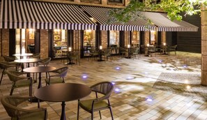 An outdoor cafe area with tables and chairs, a striped awning, and a brick wall. - Sun Street Hotel Shoreditch (London)