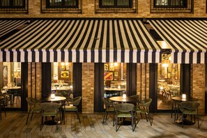 An outdoor dining area with striped awnings, brick walls, and wicker chairs. - Sun Street Hotel Shoreditch (London)