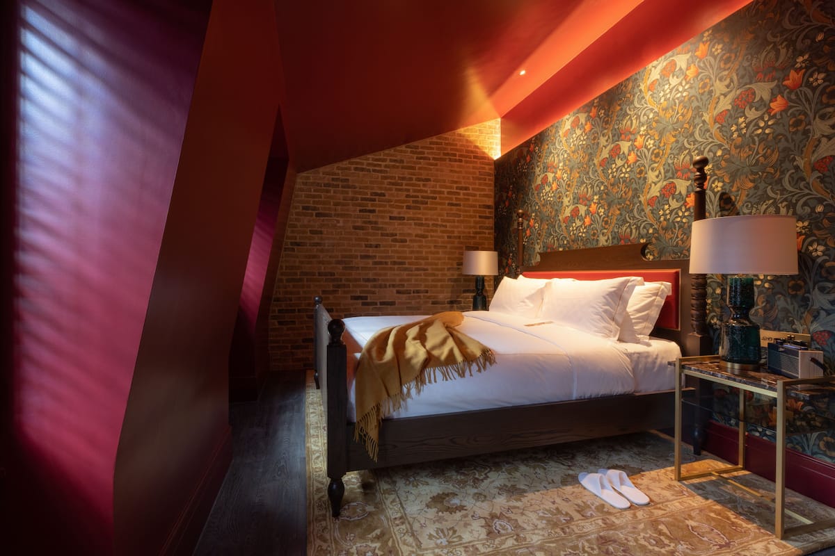 A bedroom with a brick wall, a patterned wallpaper, a bed with white linens, a nightstand with a lamp, and a pair of slippers on the floor.