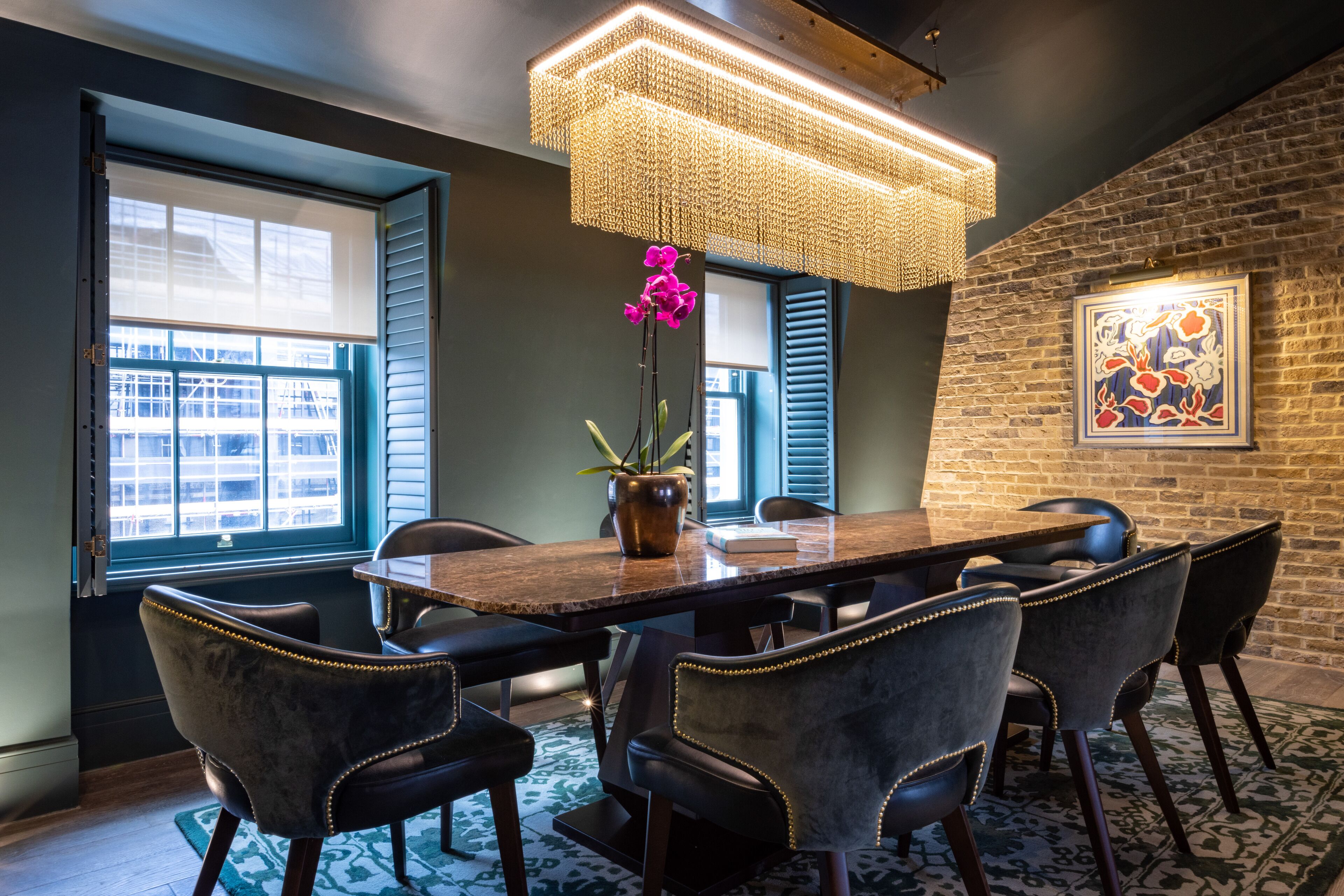 A dining area with a wooden table, black upholstered chairs, a hanging light fixture, and a potted orchid.