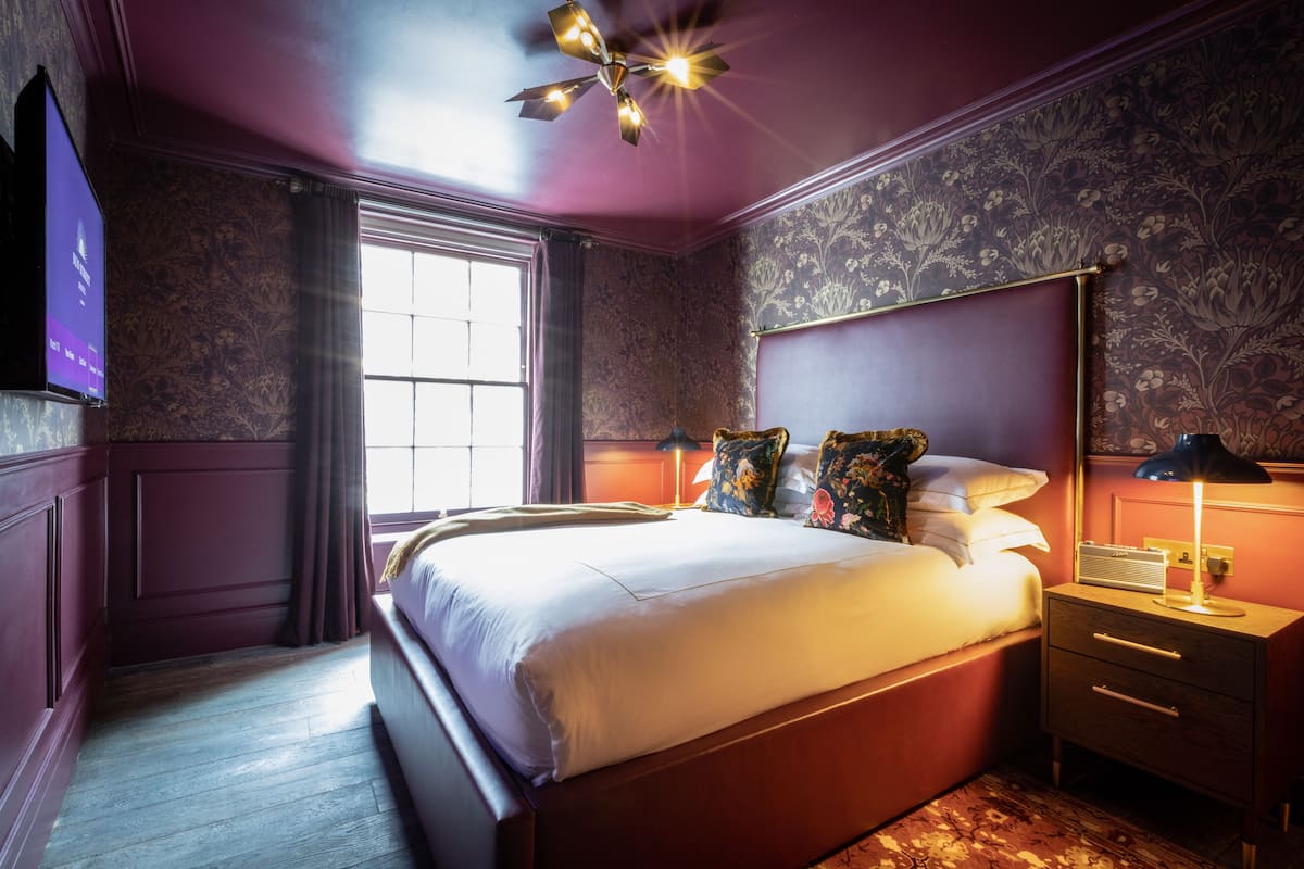 A bedroom with a large bed, a nightstand with a lamp, a dresser, and a television on the wall.