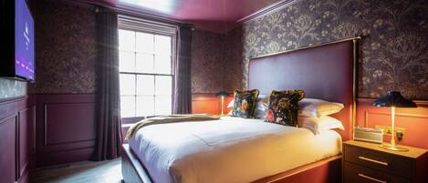 A bedroom with a large bed, a nightstand with a lamp, a dresser, and a television on the wall.