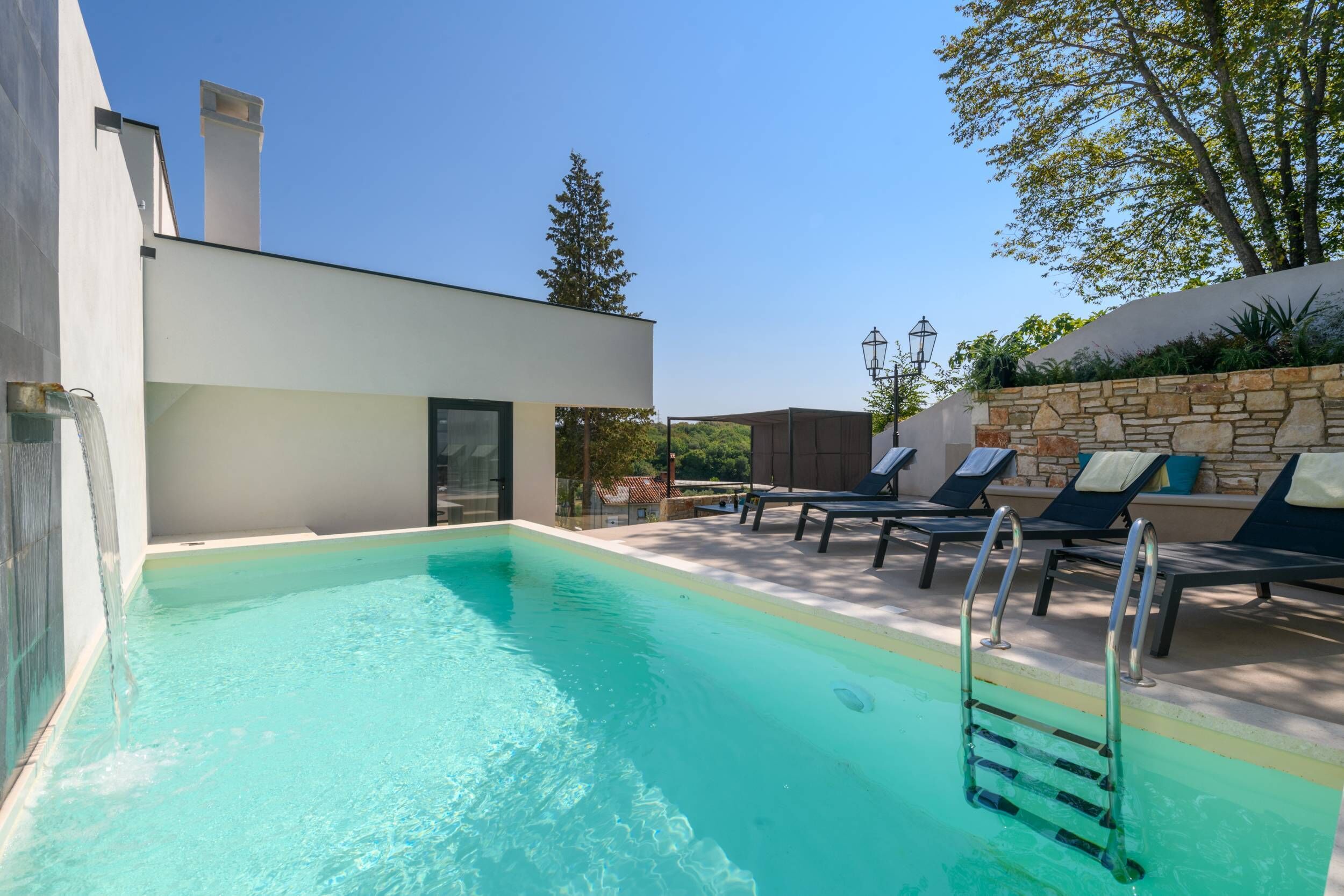 Un espace piscine extérieur moderne, doté d’une piscine à l’eau d’un bleu limpide, de chaises longues et d’un mur en pierre.