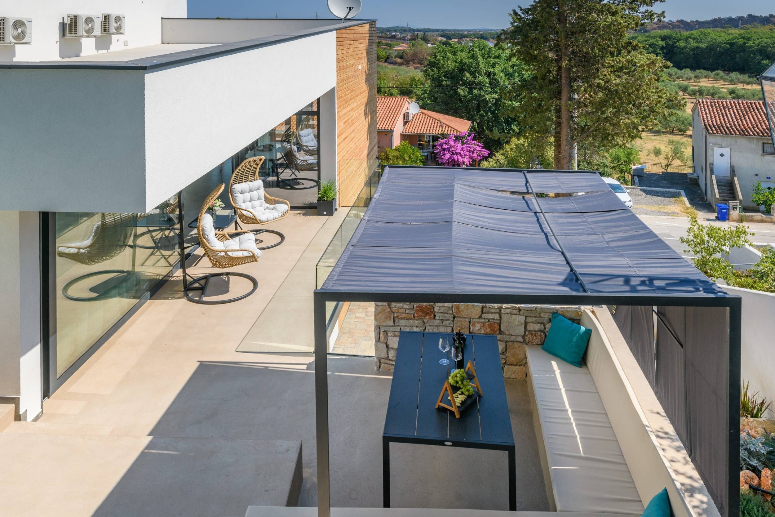 Un patio moderno al aire libre con barandilla de vidrio, muebles de mimbre y una mesa de piedra con una copa de vino y uvas.