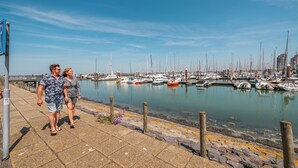 Un puerto deportivo con barcos amarrados, cielo despejado y una pareja caminando por el paseo marítimo.