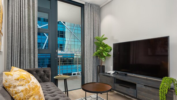A modern living room with a large flat-screen TV, a grey sofa, and a small round table. - Modern Designer Apartment in Bree Street (Cape Town)