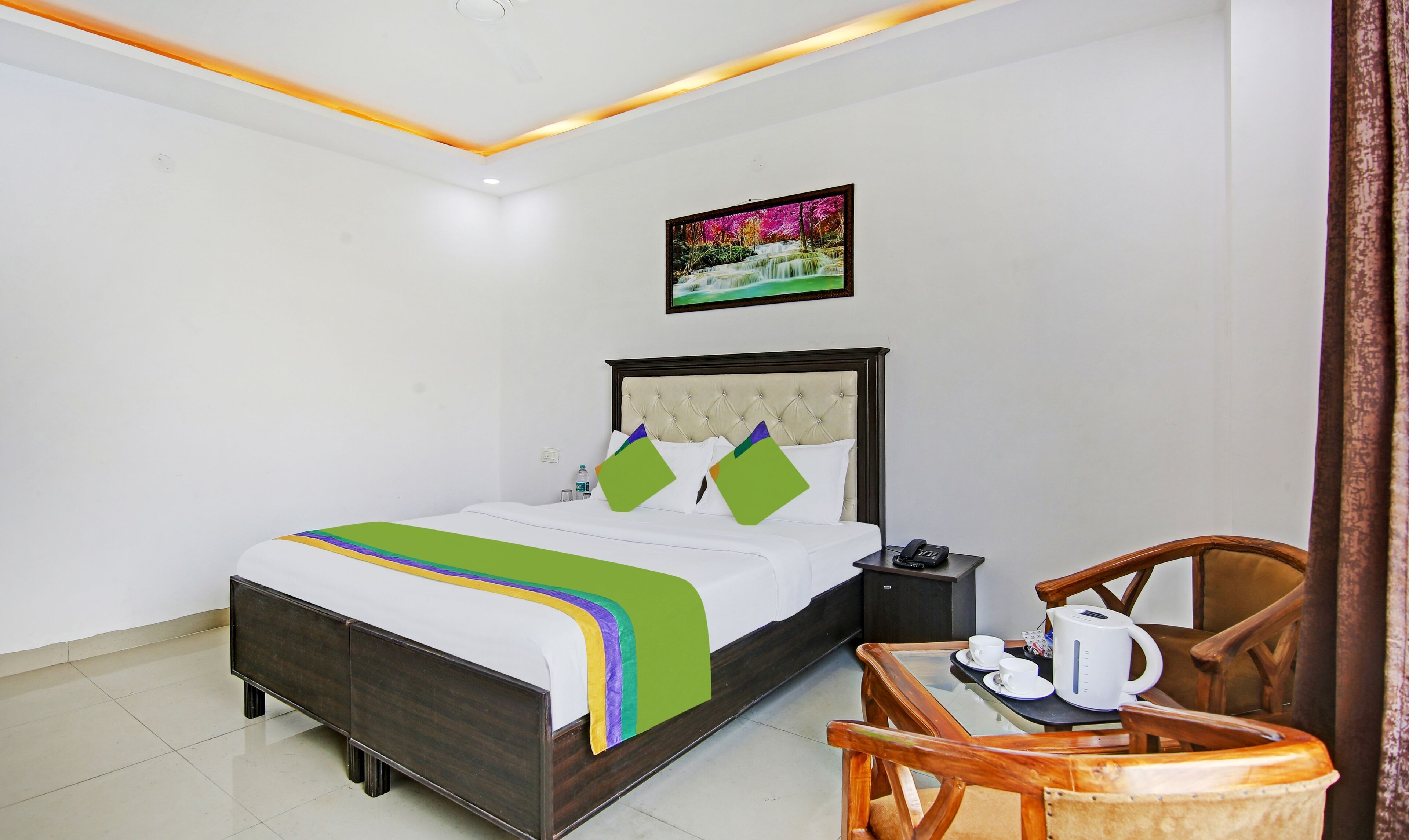 A hotel room with a bed, a bedside table with a phone, a chair, and a table with a kettle and cups.