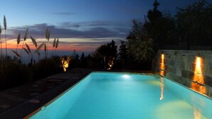 A swimming pool at dusk with a sunset view, surrounded by trees and a stone wall with lights.