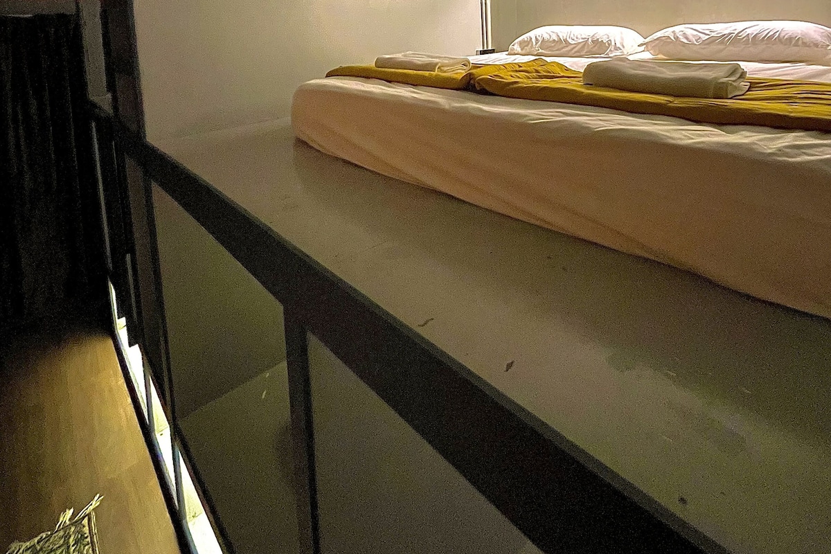 A bunk bed with a mattress and pillows, a window with a view, and a wall-mounted shelf.