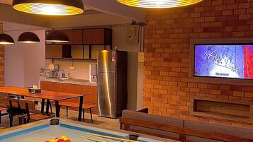 A pool table with billiard balls, a television mounted on a brick wall, and a kitchen area with a refrigerator.