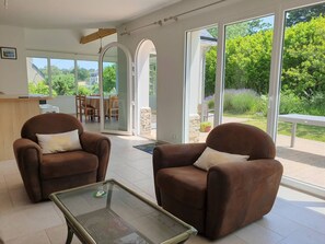 Un salon moderne avec deux fauteuils marron, une table basse en verre et une vue sur un jardin grâce à de grandes fenêtres.