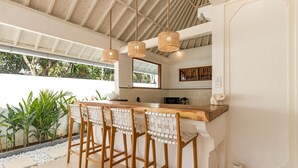 A kitchen with a bar area, wooden stools, hanging wicker lights, and a view of greenery outside.