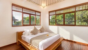 A bedroom with a wooden bed, white bedding, and a view of greenery through wooden blinds.