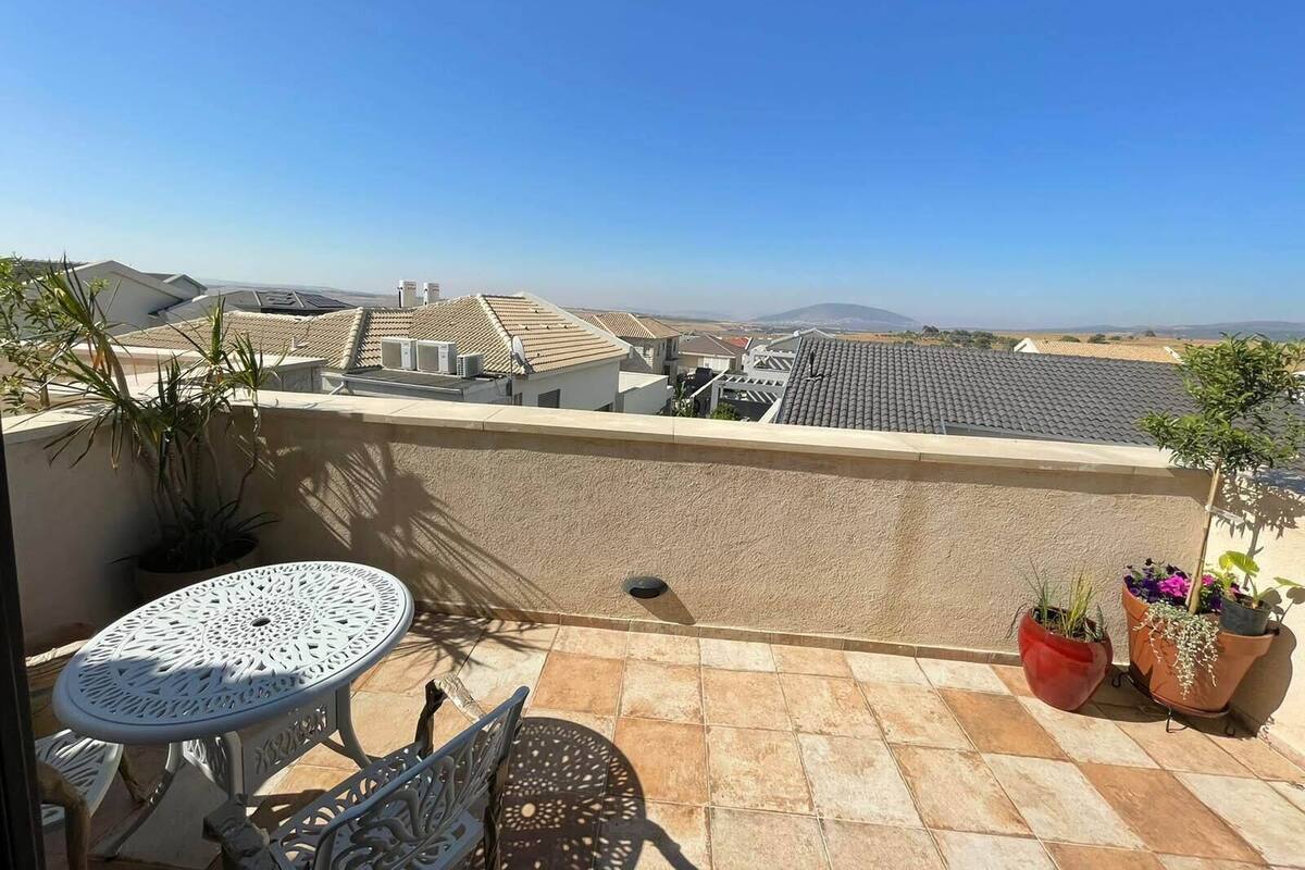 A balcony with a table and chairs, potted plants, and a view of neighboring buildings and hills.