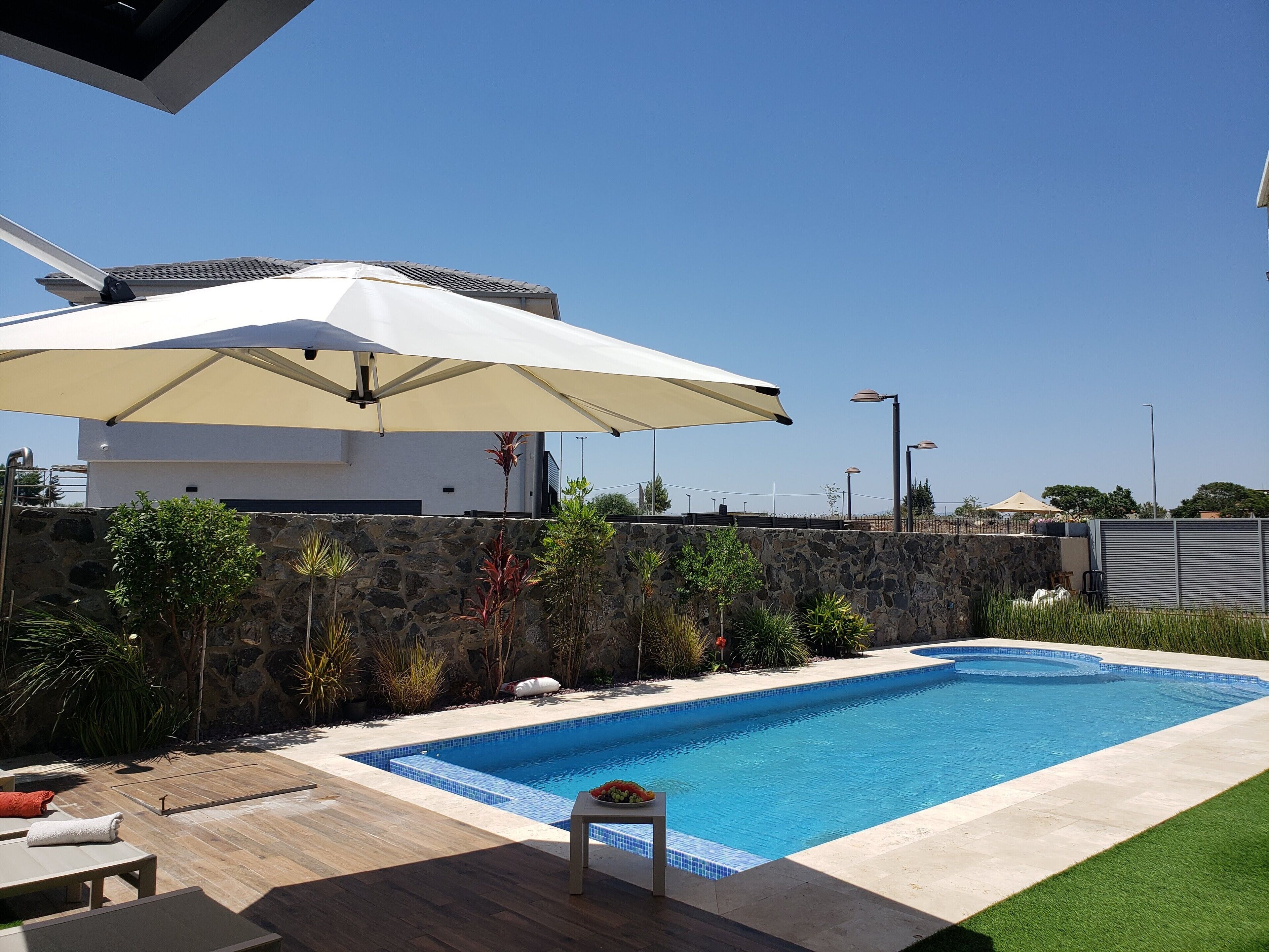 En swimmingpool med en stenvæg, en træterrasse og en hvid parasol.