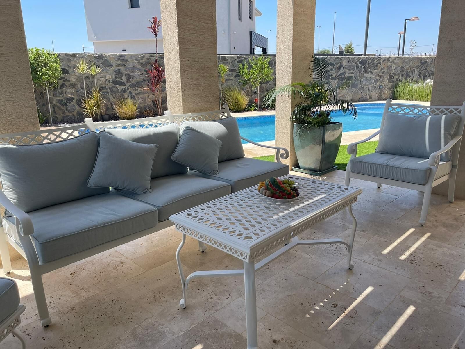 A patio with a white sofa, two white chairs, a white table, and a pool in the background.