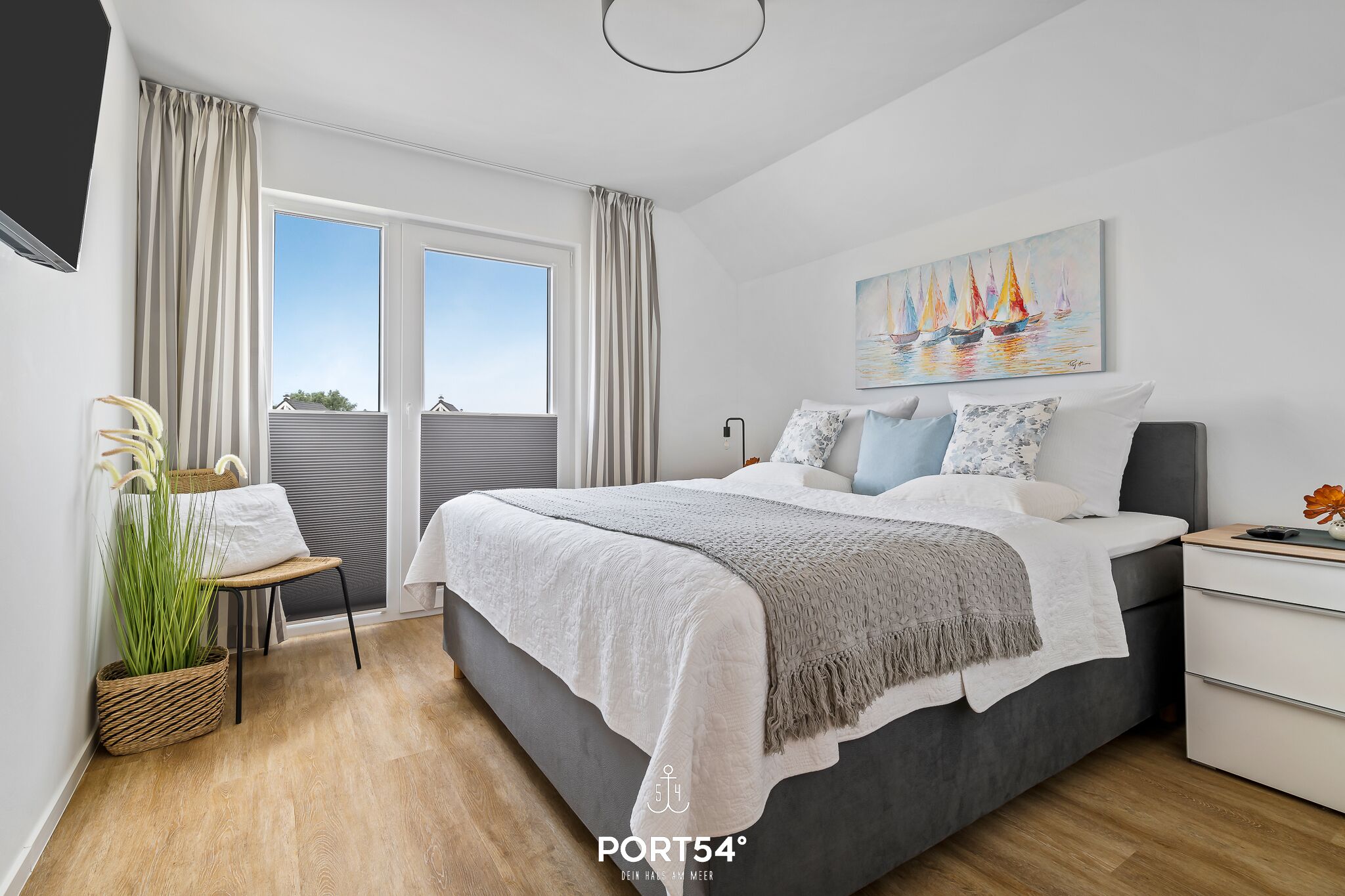 A modern bedroom with a large bed, a bedside table, a window with curtains, a painting on the wall, and a plant.