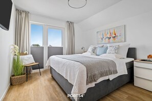 A modern bedroom with a large bed, a bedside table, a window with curtains, a painting on the wall, and a plant.
