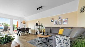 A modern living room with a sofa, coffee table, and chairs. There is a dining area with a table and chairs. The room has large windows and a view of the outside.