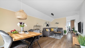 Un salon moderne avec une table à manger en bois, des chaises à revêtement gris, un canapé gris avec des coussins jaunes, un lampadaire noir et un téléviseur placé à droite.