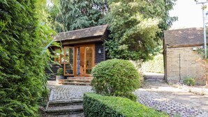 A rustic cabin with a stone pathway leading to a wooden door, surrounded by dense greenery and a brick wall. - Willow Cottage (Dartford)